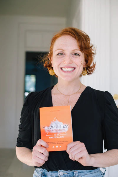Lara Hocheiser holding her 2021 book 'Mindfulness for Beginners in Ten Minutes a Day' smiling. She had red hair, gold tree of life earnings, and is smiling. 